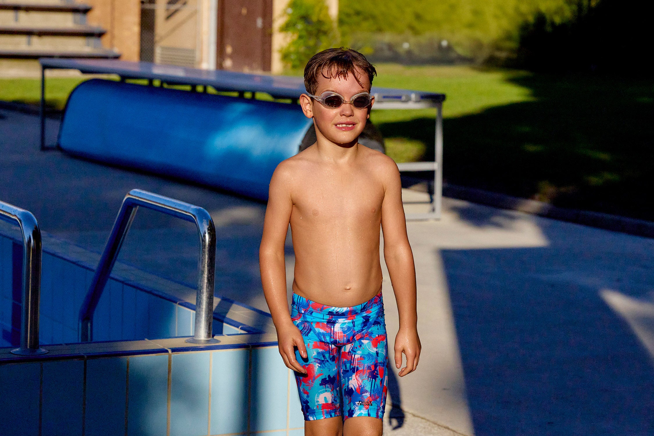 Young boy in swim trunks and sunglasses standing by a pool.