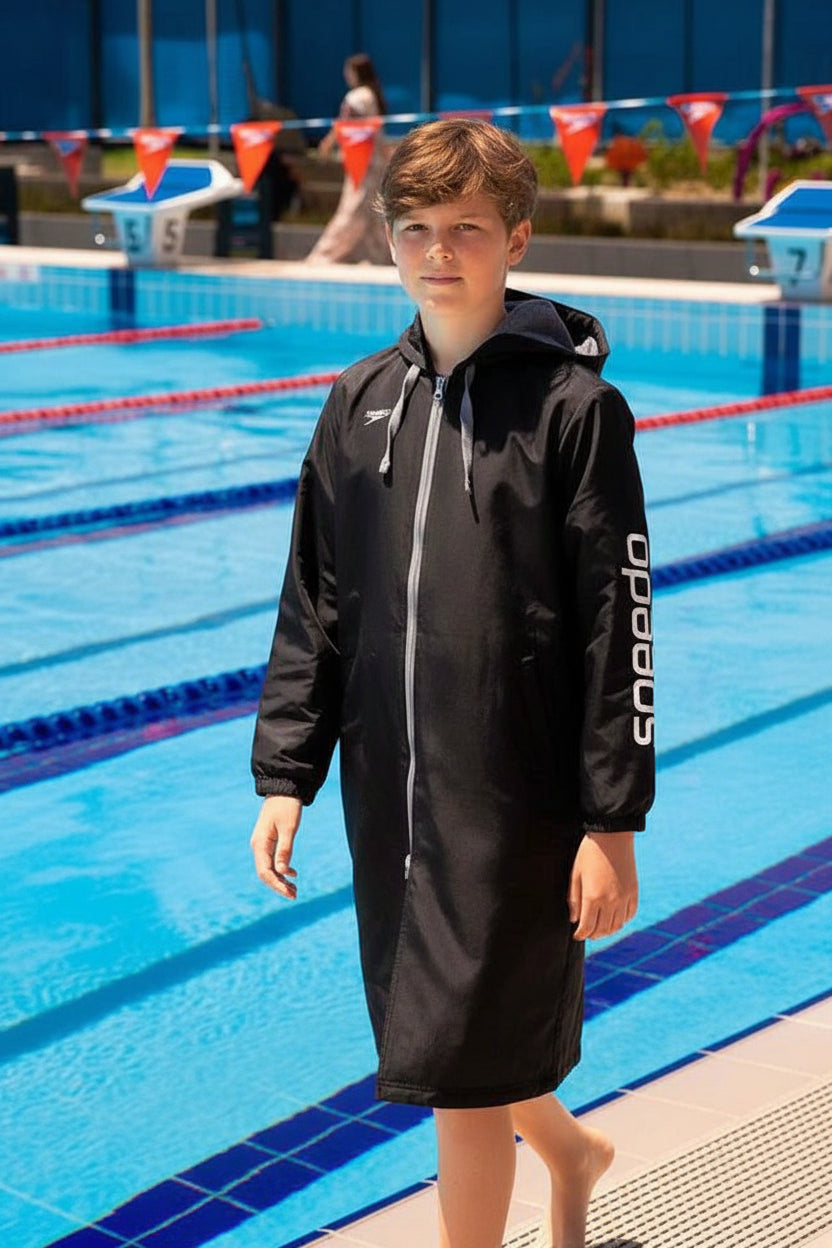 A young boy walking in front of a swimming pool wearing a black Speedo hooded swim towel jacket. The jacket features a full front zipper, a drawstring hood, and the brand name 'Speedo' printed vertically in white on the left sleeve.