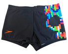 Black athletic swim trunks with a bright multicolor geometric pattern on the right leg and an orange Speedo logo on the left leg, laid flat on a wood-grain surface.