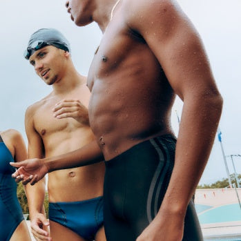 Two swimmers standing by a pool, wearing swimsuits and swim caps.